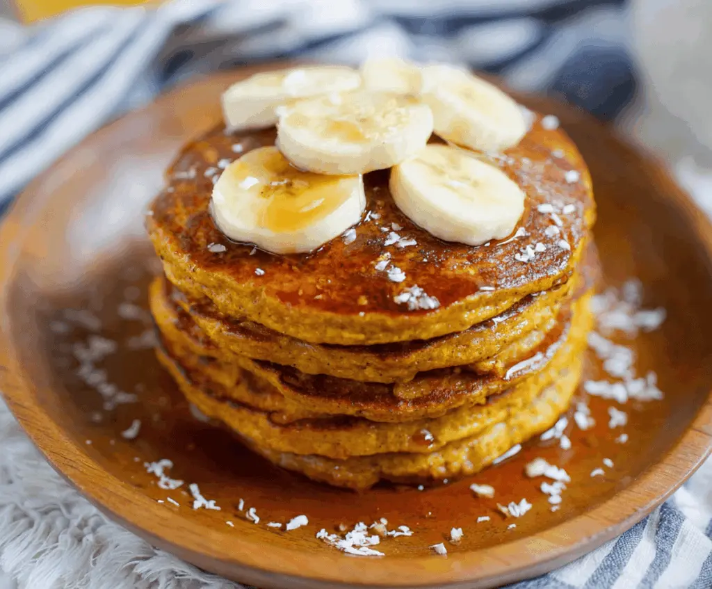 Delicious homemade pumpkin banana pancakes topped with whipped cream and cinnamon, served on a white plate with fresh pumpkin and bananas in the background.