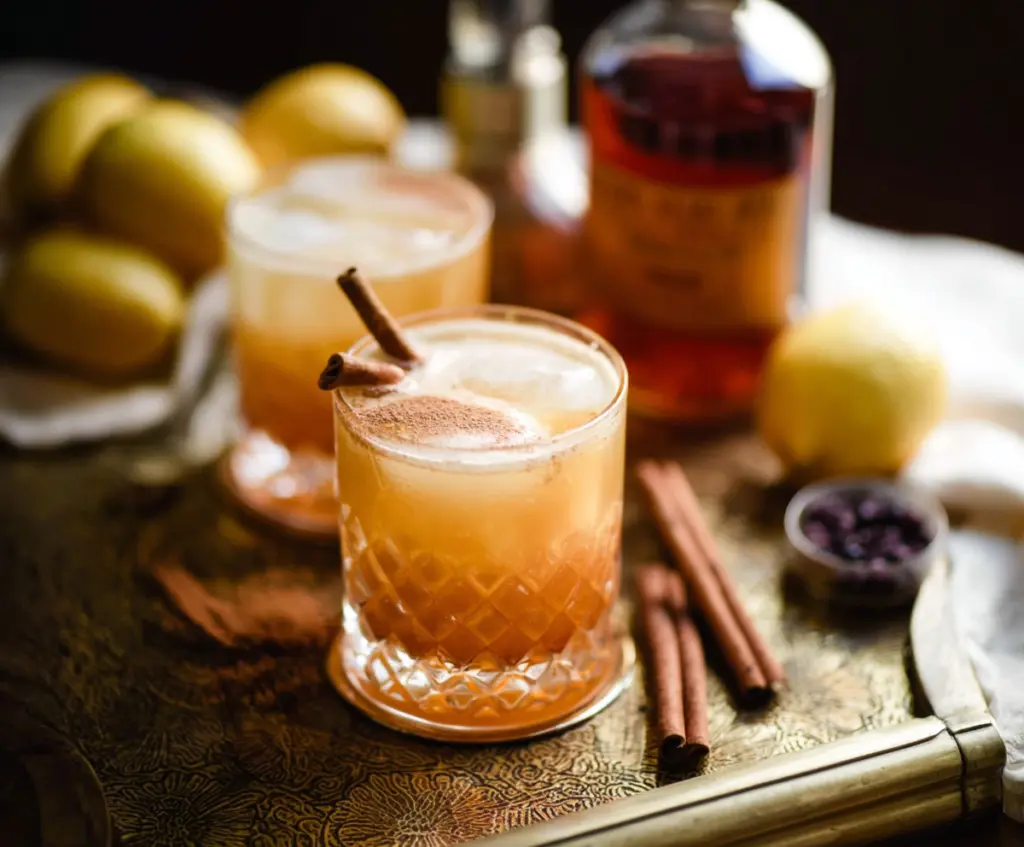 A cocktail glass filled with a Cinnamon Maple Whiskey Sour garnished with a cinnamon stick and a slice of maple syrup-infused lemon.